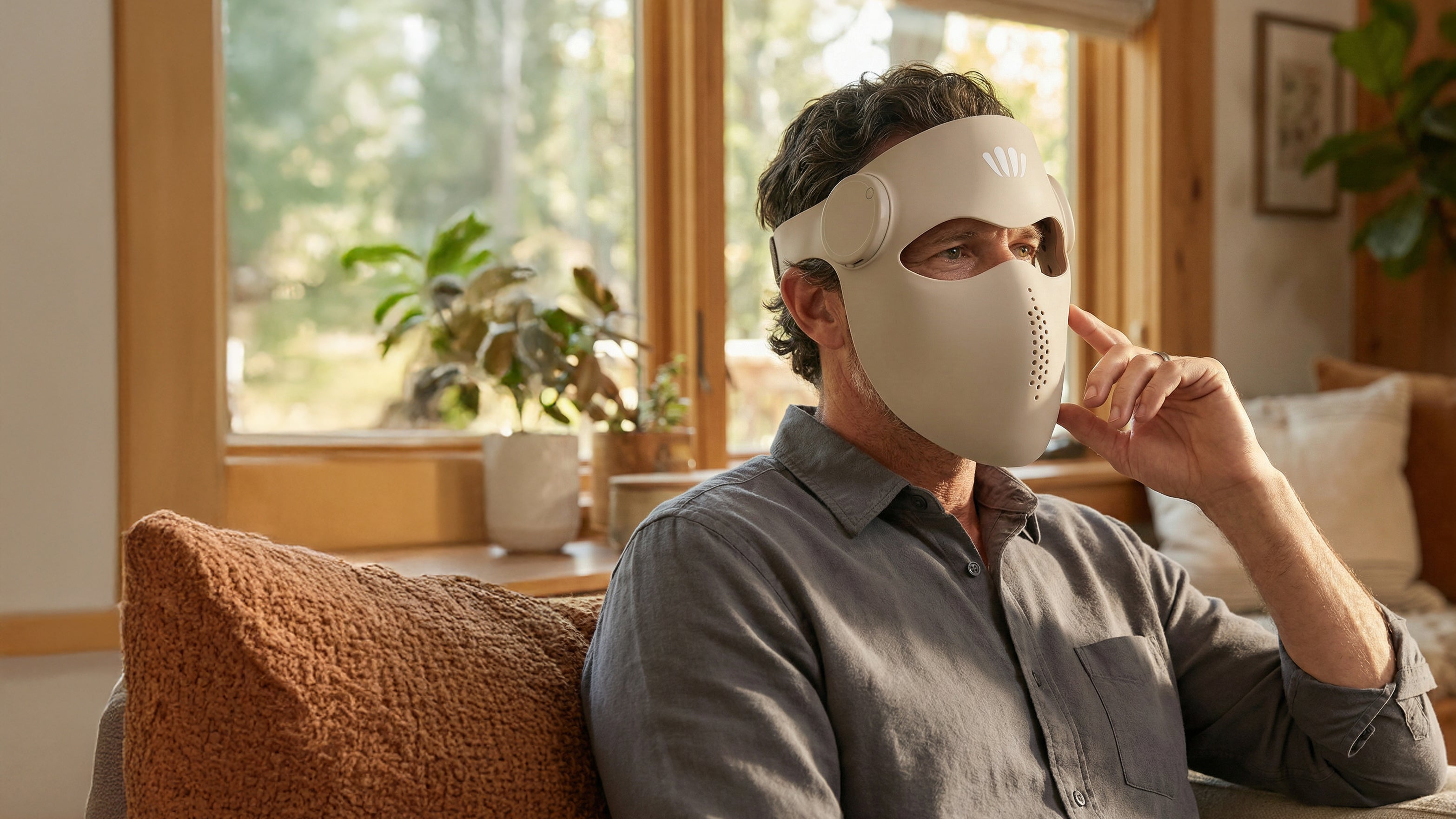 Person wearing a beige silicone red light therapy facemask in a cozy living room with large windows.