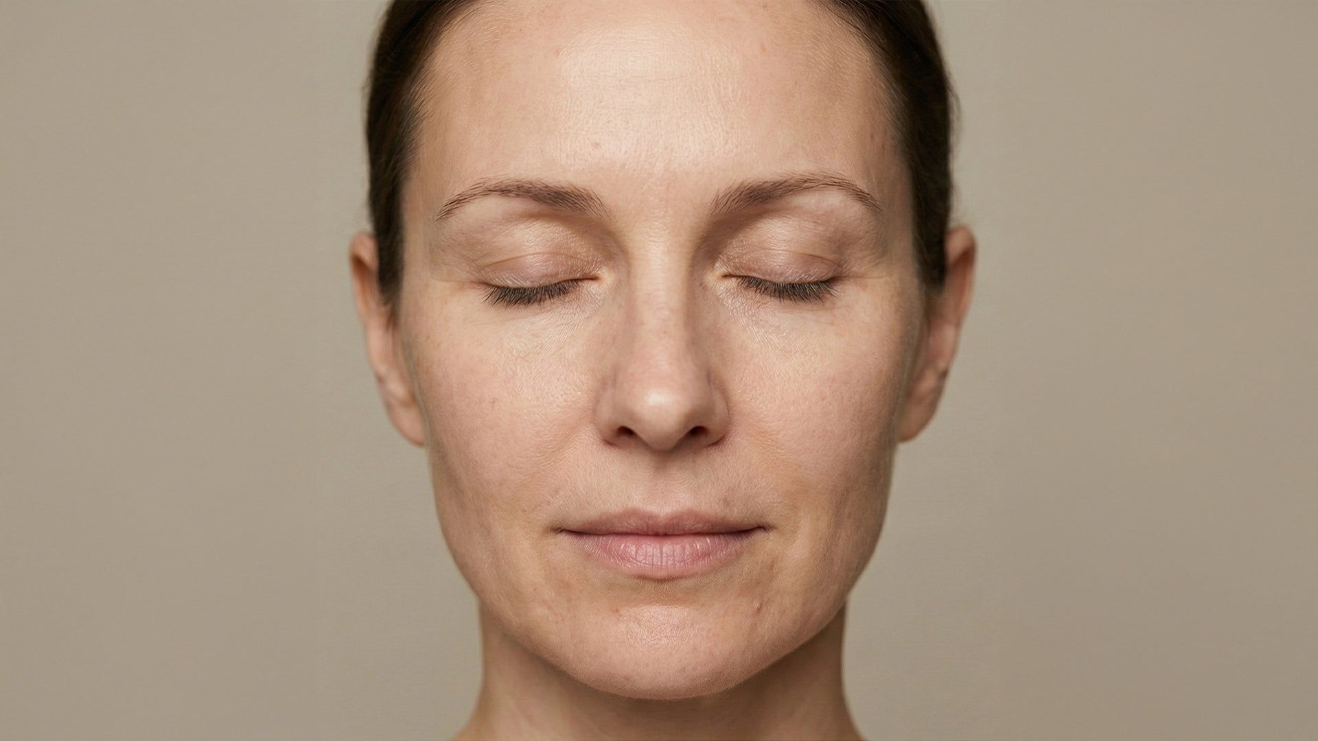Close-up of a woman with her eyes closed against a beige background before red light therapy face treatment